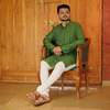 Man in a green kurta sitting on a wooden chair against a wooden door,wearing brown kapshi kolhapuri chappal