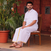 Man in traditional white attire sitting on a wooden chair with plants and a red wall in the background